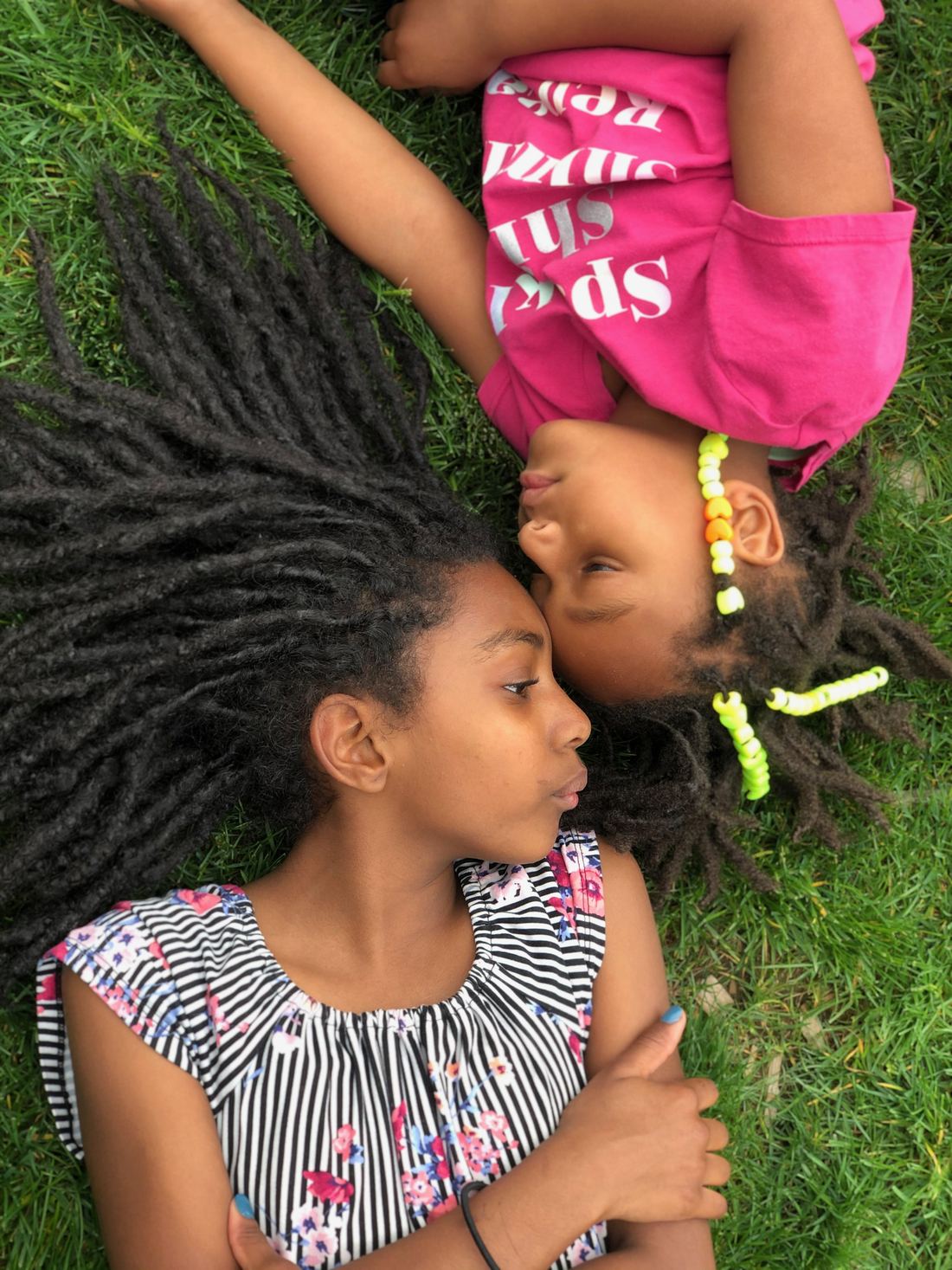 Two girls lying head to head on the grass, one with long locs, one with beaded locs